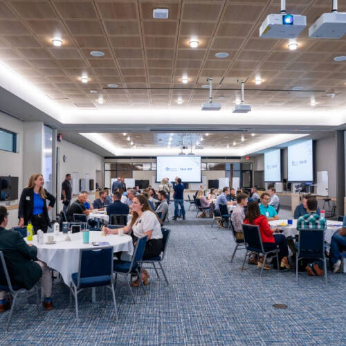 People seated in small groups at round tables during a professional event in a large, modern conference room with presentation screens and ceiling projectors.
